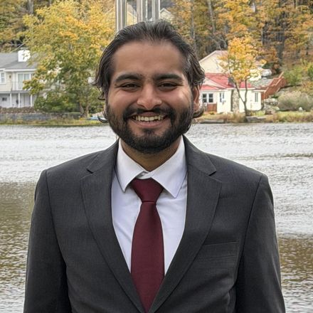 Nick Sawhney, smiling, in a grey suit and dark red tie, standing in front of a lake with autumn trees behind him.